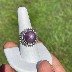 May include: A silver ring with a purple star ruby gemstone set in a silver sunburst design.
