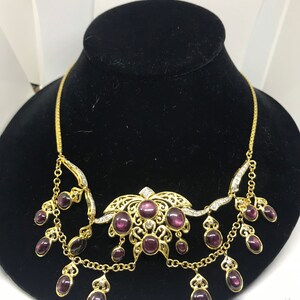 May include: A gold necklace with a large, ornate pendant featuring a floral design and multiple purple stones. The pendant is surrounded by smaller purple stones that dangle from the chain.