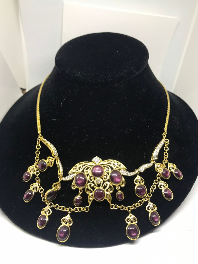May include: A gold necklace with a large, ornate pendant featuring a floral design and multiple purple stones. The pendant is surrounded by smaller purple stones that dangle from the chain.
