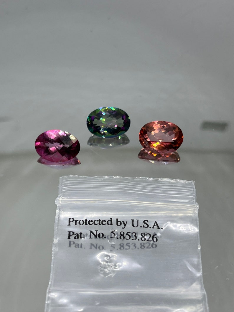 Puede incluir: Tres piedras preciosas de forma ovalada, una verde, una rosa y una melocot&oacute;n, se muestran sobre una superficie blanca. Una peque&ntilde;a bolsa de pl&aacute;stico con el texto "Protected by U.S.A. Pat. No. 5,853,826" se encuentra en la esquina inferior derecha de la imagen.