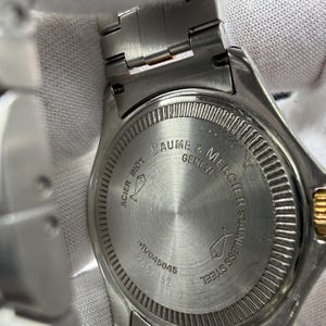 May include: Close-up of a stainless steel Baume & Mercier watch. The watch back features the brand name, model number, and the words "ACIER INOX" and "STAINLESS STEEL". The watch band is silver.
