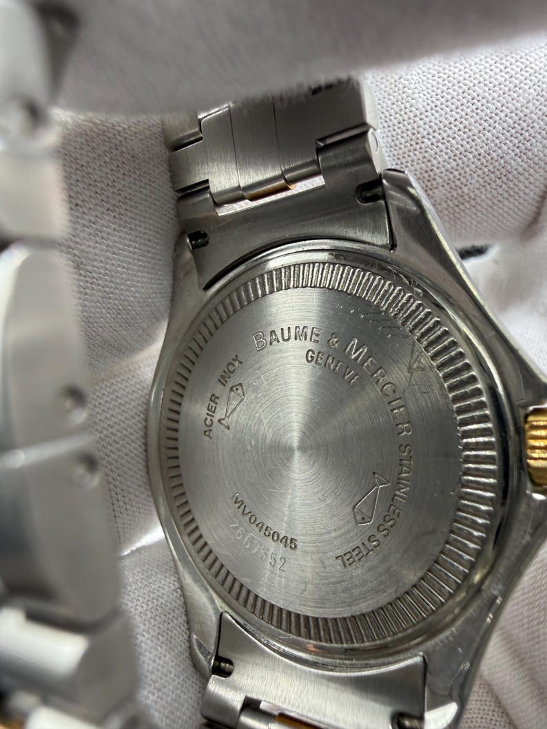 May include: Close-up of a stainless steel Baume & Mercier watch. The watch back features the brand name, model number, and the words "ACIER INOX" and "STAINLESS STEEL". The watch band is silver.