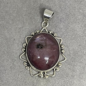 Puede incluir: Un colgante de plata con una piedra preciosa grande, ovalada, de color púrpura oscuro engastada en un diseño de estrella.