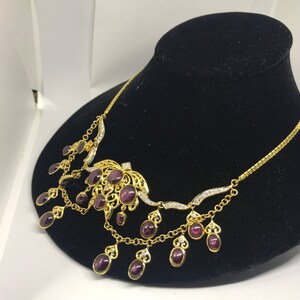 May include: A gold necklace with a large purple gemstone pendant and smaller purple gemstones dangling from the chain. The necklace is on a black velvet display stand.