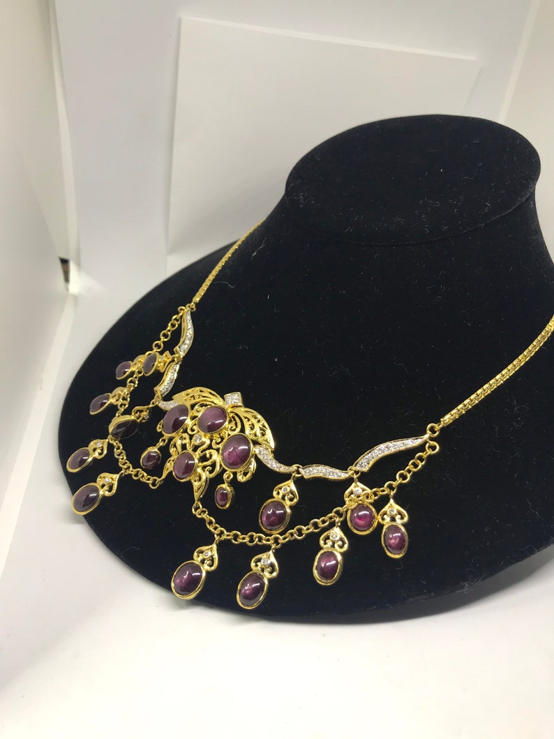 May include: A gold necklace with a large purple gemstone pendant and smaller purple gemstones dangling from the chain. The necklace is on a black velvet display stand.