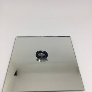 Puede incluir: Una piedra preciosa de color p&uacute;rpura oscuro y verde con forma ovalada facetada, expuesta en un espejo plateado.