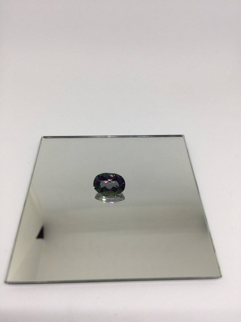 Puede incluir: Una piedra preciosa de color p&uacute;rpura oscuro y verde con forma ovalada facetada, expuesta en un espejo plateado.
