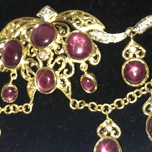 May include: A gold necklace with a pendant featuring a cluster of red gemstones and a delicate filigree design. The necklace has a chain with a dangling pendant featuring a single red gemstone.