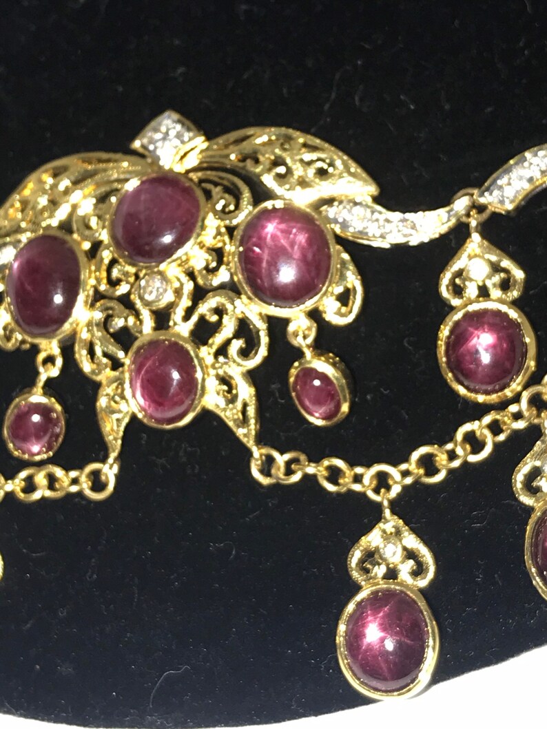 May include: A gold necklace with a pendant featuring a cluster of red gemstones and a delicate filigree design. The necklace has a chain with a dangling pendant featuring a single red gemstone.