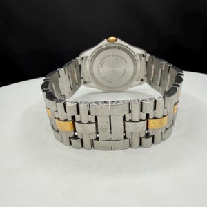 May include: A stainless steel wristwatch with gold-toned accents. The watch band is a combination of silver and gold links. The watch face is not visible, but the back of the watch is visible with text. The watch is on a white surface.