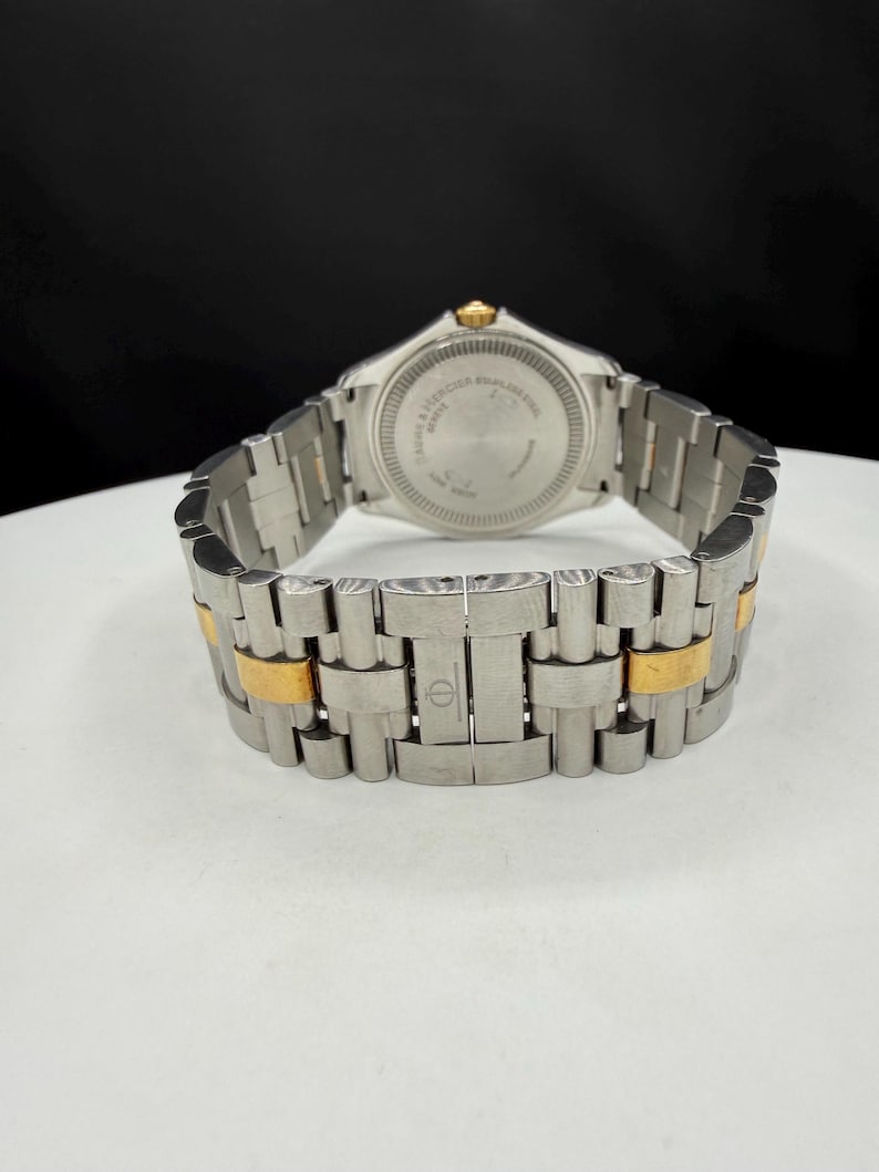May include: A stainless steel wristwatch with gold-toned accents. The watch band is a combination of silver and gold links. The watch face is not visible, but the back of the watch is visible with text. The watch is on a white surface.