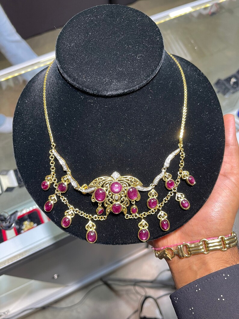 May include: A gold necklace with a delicate chain and a pendant featuring a cluster of pink gemstones surrounded by smaller diamonds. The pendant is connected to a second chain with dangling pink gemstones.