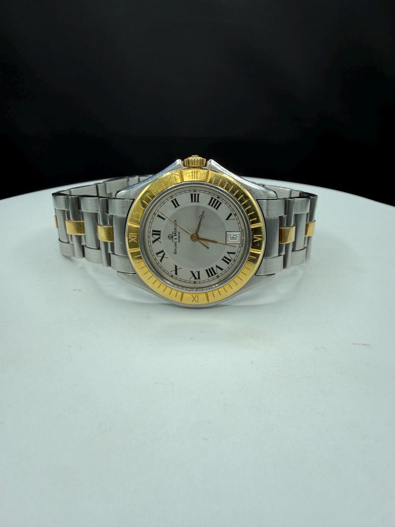 May include: A silver and gold-toned wristwatch with a silver dial and gold Roman numeral hour markers. The watch has a date window at the 3 o'clock position and a two-tone stainless steel and gold band. The watch is displayed on a white surface.