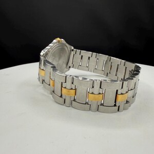 May include: A stainless steel watch with gold-tone accents on the band. The watch face is silver with a gold-tone bezel. The watch is displayed on a white surface with a black background.