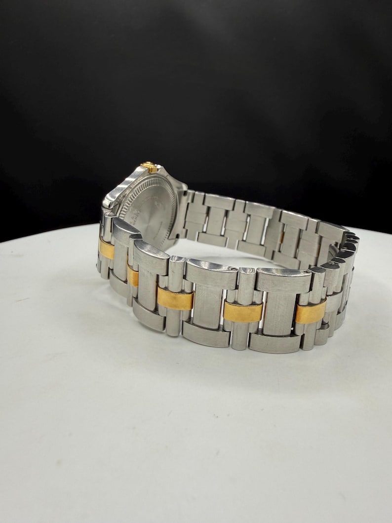 May include: A stainless steel watch with gold-tone accents on the band. The watch face is silver with a gold-tone bezel. The watch is displayed on a white surface with a black background.