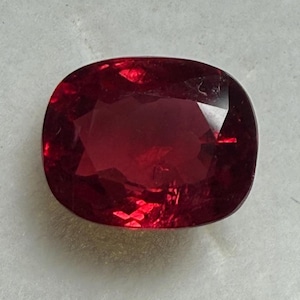 Puede incluir: Una piedra preciosa de color rojo intenso con talla cojín. La piedra preciosa tiene forma ovalada y una superficie lisa y pulida.