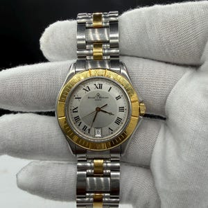 May include: A Baume & Mercier Geneve watch with a silver face, gold bezel, and silver and gold band. The watch has Roman numeral hour markers and a date display at the 3 o'clock position. The watch band is made of stainless steel and gold links.
