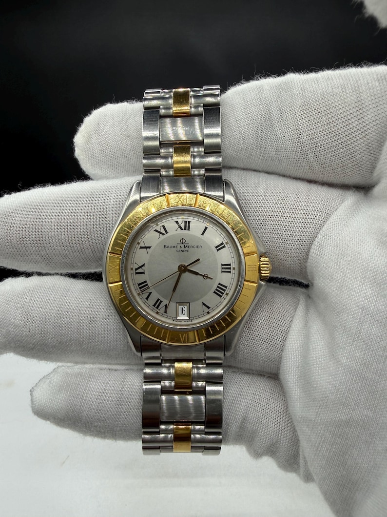 May include: A Baume & Mercier Geneve watch with a silver face, gold bezel, and silver and gold band. The watch has Roman numeral hour markers and a date display at the 3 o'clock position. The watch band is made of stainless steel and gold links.