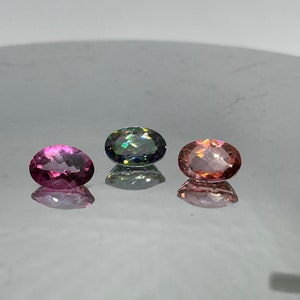 Puede incluir: Tres piedras preciosas de forma ovalada, una rosa, una verde con brillo iridiscente y una melocot&oacute;n, dispuestas sobre una superficie blanca.