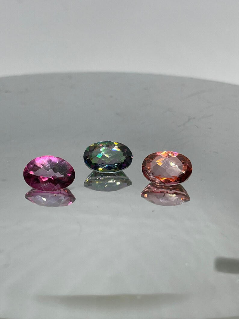 Puede incluir: Tres piedras preciosas de forma ovalada, una rosa, una verde con brillo iridiscente y una melocot&oacute;n, dispuestas sobre una superficie blanca.