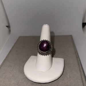 May include: A silver ring with a purple star ruby gemstone set in a bezel setting. The ring has a decorative border of small silver beads.
