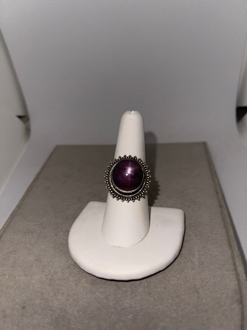 May include: A silver ring with a purple star ruby gemstone set in a bezel setting. The ring has a decorative border of small silver beads.