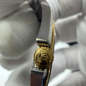 May include: Close-up of a luxury wristwatch. The watch features a gold crown with a logo and a silver and gold band. The watch is set against a white background.