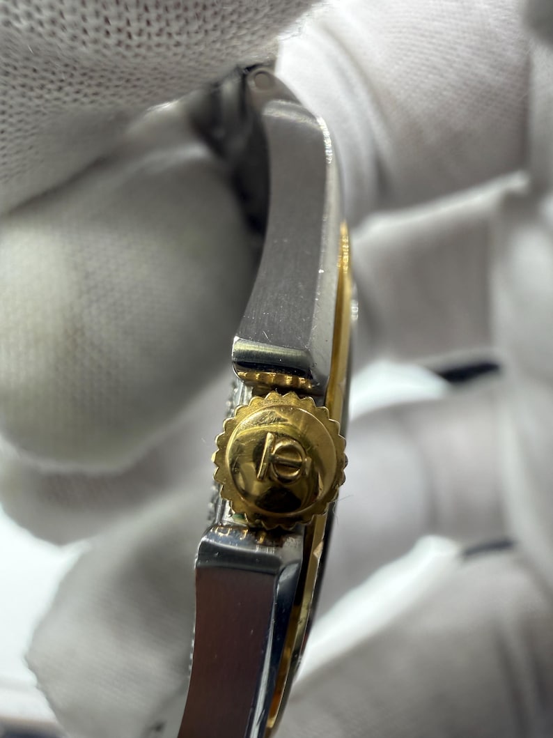 May include: Close-up of a luxury wristwatch. The watch features a gold crown with a logo and a silver and gold band. The watch is set against a white background.