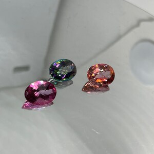 Puede incluir: Tres piedras preciosas ovaladas, una verde, una rosa y una roja, se muestran sobre una superficie reflectante.