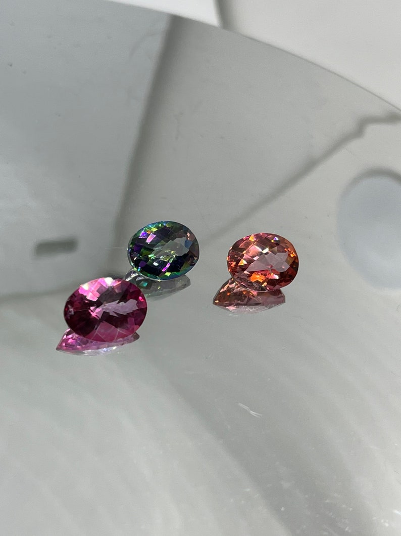Puede incluir: Tres piedras preciosas ovaladas, una verde, una rosa y una roja, se muestran sobre una superficie reflectante.