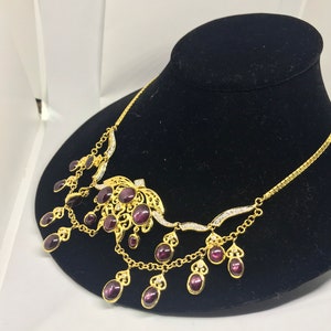 May include: A gold necklace with a purple stone centerpiece and dangling purple stones. The necklace has a delicate chain and intricate detailing.