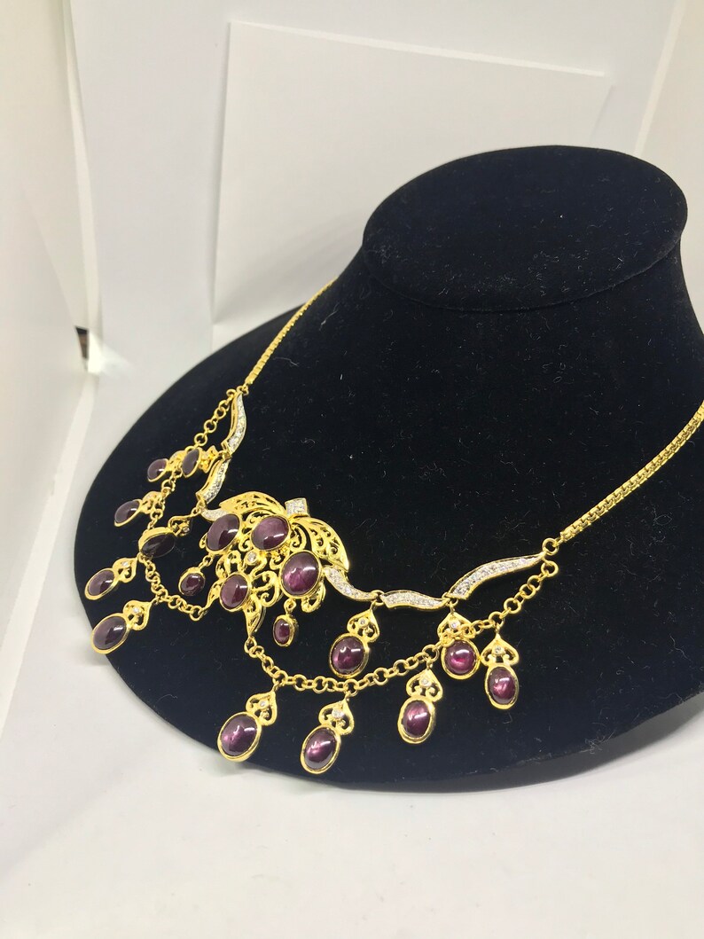 May include: A gold necklace with a purple stone centerpiece and dangling purple stones. The necklace has a delicate chain and intricate detailing.