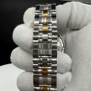 May include: Close-up of a stainless steel watch band with gold-colored accents. The band is composed of multiple rectangular links, alternating between silver and gold tones. The watch face is partially visible.