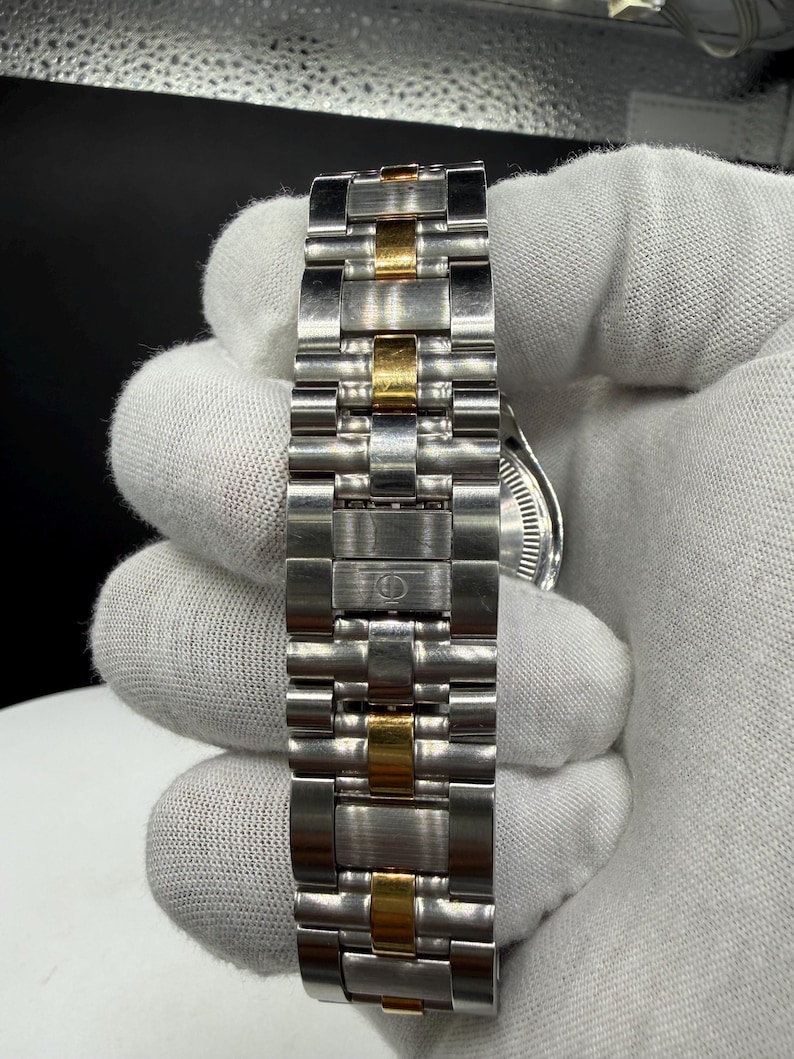 May include: Close-up of a stainless steel watch band with gold-colored accents. The band is composed of multiple rectangular links, alternating between silver and gold tones. The watch face is partially visible.