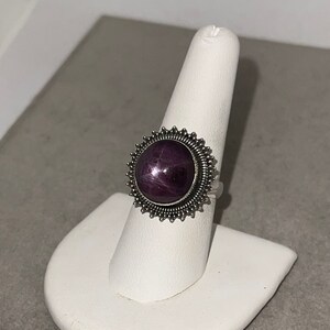 May include: A silver ring with a purple star ruby gemstone set in a circular, starburst-shaped bezel.