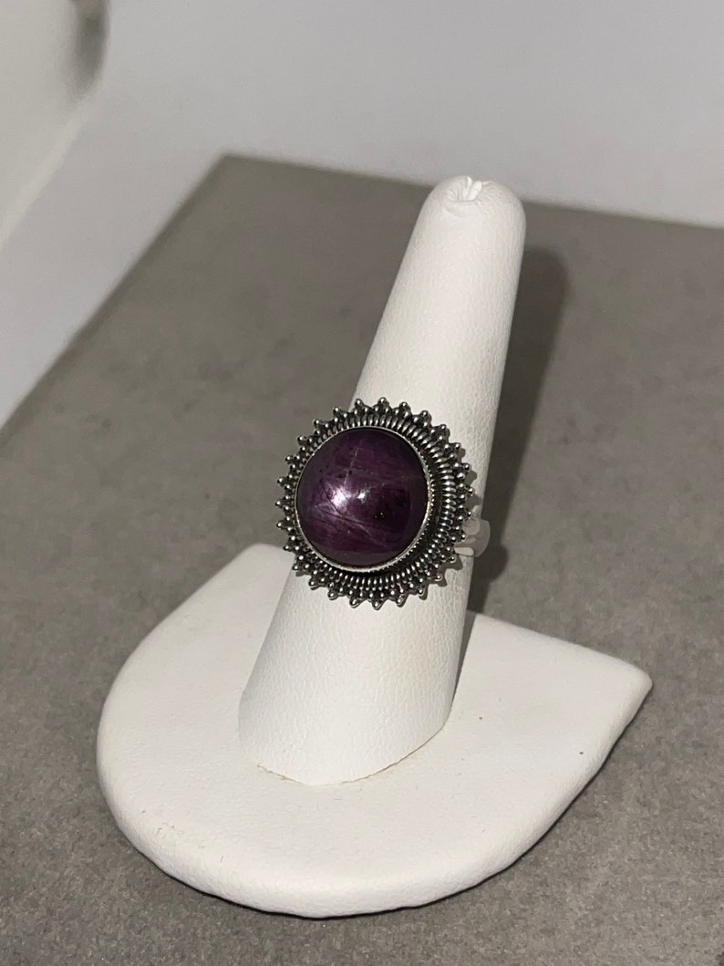 May include: A silver ring with a purple star ruby gemstone set in a circular, starburst-shaped bezel.