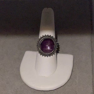 May include: A silver ring with a purple gemstone set in a decorative, circular bezel. The ring is displayed on a white ring stand.