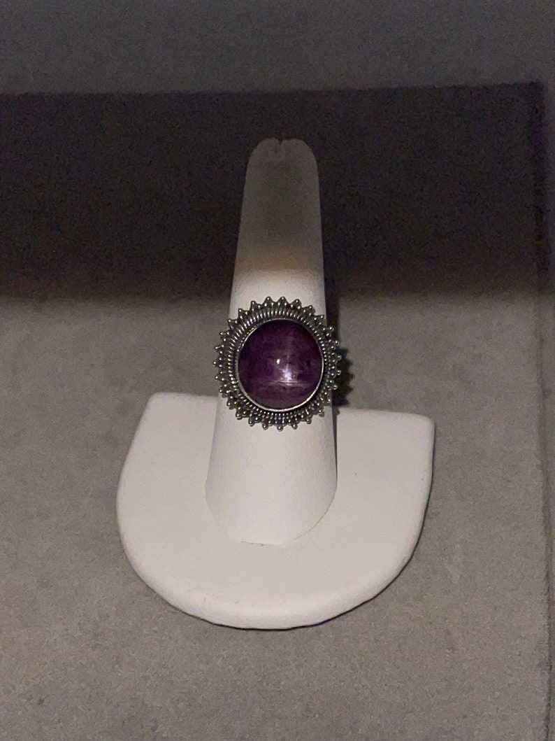 May include: A silver ring with a purple gemstone set in a decorative, circular bezel. The ring is displayed on a white ring stand.