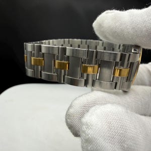 May include: A stainless steel watch band with gold-colored accents. The band is composed of rectangular links, alternating between silver and gold. The watch band is shown against a dark background.