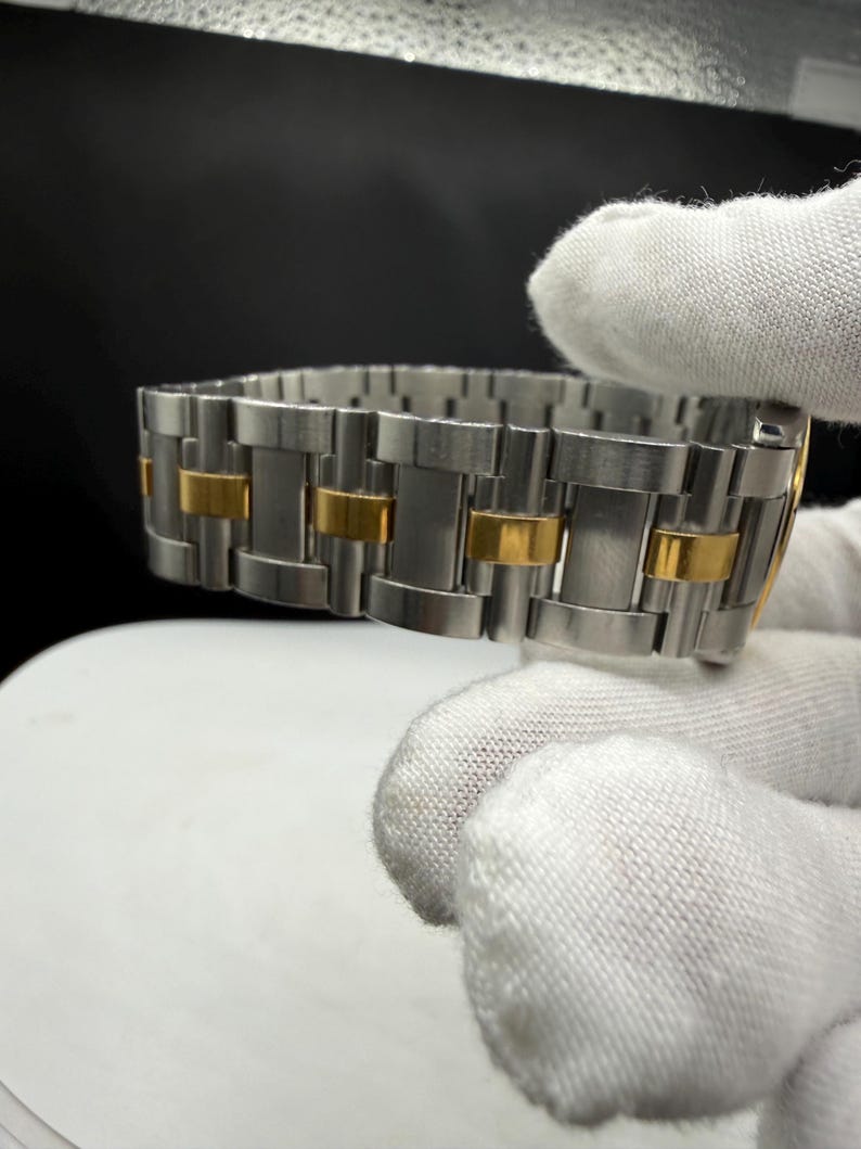 May include: A stainless steel watch band with gold-colored accents. The band is composed of rectangular links, alternating between silver and gold. The watch band is shown against a dark background.