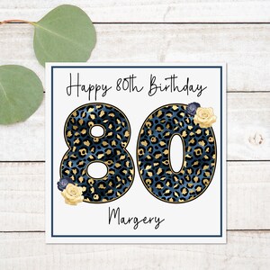 Puede incluir: Una tarjeta blanca con un borde azul y el texto "Happy 80th Birthday" en letra cursiva negra. El n&uacute;mero 80 est&aacute; en letras grandes, negritas, de estampado de leopardo azul y dorado. El nombre "Margery" est&aacute; escrito en letra cursiva negra debajo del n&uacute;mero 80.