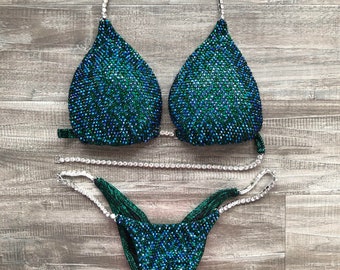 May include: A green and blue sequined bikini with crystal embellishments. The bikini top has a halter style strap and the bottom has a high-cut leg.