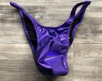 Può includere: Uno slip bikini a taglio alto in tessuto viola metallizzato con una finitura scintillante. Il capo presenta un esterno viola a tinta unita con una fodera nera a contrasto visibile lungo le aperture delle gambe e i bordi della vita.