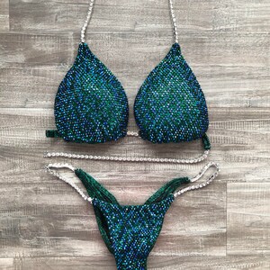 May include: A green and blue sequined bikini with crystal embellishments. The bikini top has a halter style strap and the bottom has a high-cut leg.