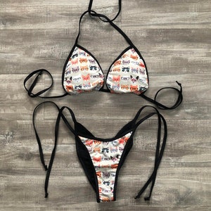 May include: A black bikini with a white background featuring a repeating pattern of cats wearing glasses. The bikini top is a triangle style with adjustable straps. The bikini bottom is a high-cut style with adjustable ties on the sides.