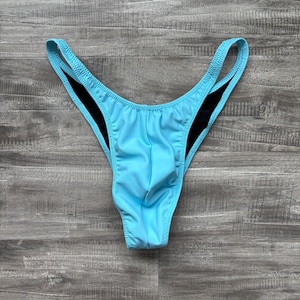 May include: Light blue thong-style swimwear with black trim. The swimsuit has a high-cut leg and a narrow waistband. The fabric appears smooth and slightly shiny. The background is a weathered wood surface.
