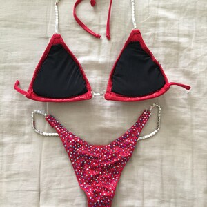 May include: Red and black bikini with crystal embellishments. The top has a halter style with a black lining. The bottom is a high-cut style with a full back.