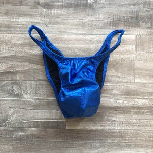 May include: A pair of high-cut posing trunks in a vibrant, metallic blue fabric with a subtle, reflective dot pattern. The garment features a contoured front pouch, thin side straps, and a solid black interior lining for structure and comfort.