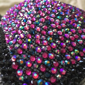 May include: Close-up of a black fabric covered with iridescent pink, blue, and black rhinestones. The rhinestones are arranged in a pattern and create a sparkling effect.