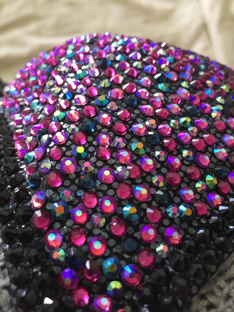 May include: Close-up of a black fabric covered with iridescent pink, blue, and black rhinestones. The rhinestones are arranged in a pattern and create a sparkling effect.